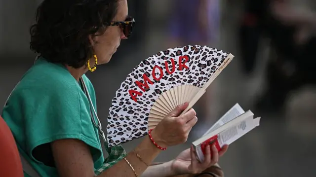 A passenger uses a fan, which has the word Amour written on it, to cool down as she waits for a train at the Saint-Jean train station