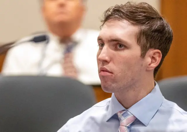 Tyler Robinson sits in court wearing a light blur shirt and a pink and blur tie