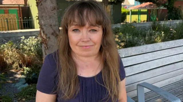 A woman with long brown hair, wearing a navy top. She's sat on a bench with trees and shrubs behind her.