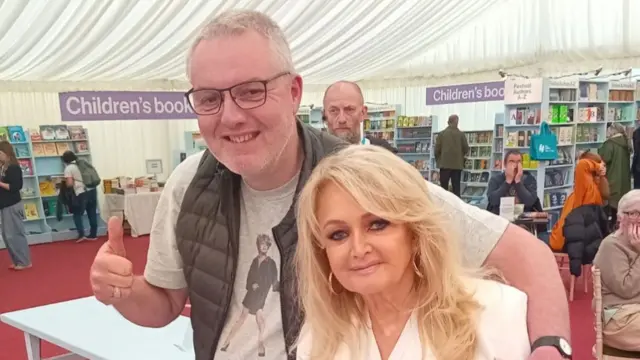 A grey-haired man with glasses wearing a grey t shirt and black gilet. He has one hanfd in a thumbs up, and the other arm around a glamorous blonde woman wearing a white blouse and big gold hoop earrings. They are both smiling and looking at the camera. Behind them are bookshelves full of books and people browsing them.