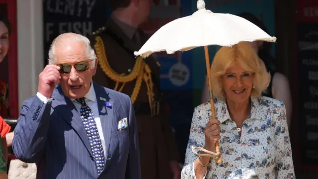 King Charles on the left wearing a striped suit and sunglasses, with Queen Camilla holding a pale umbrella to protect from the sun.