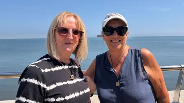 Two middle-aged blonde women. One on the left wears a striped blue and white top and sungless, the one on the right wears a navy linen vest, sunglasses and a white cap. They are stood in front of a barrier with the sea behind them and smiling.