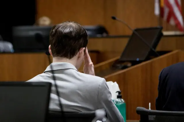 A man with brown hair and a light grey suit faces away from the camera in court