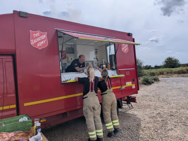 A red Salvation Army mobile canteen. A man is leaning out of its hatch and serving two firefighters, who are reaching up to receive drinks.