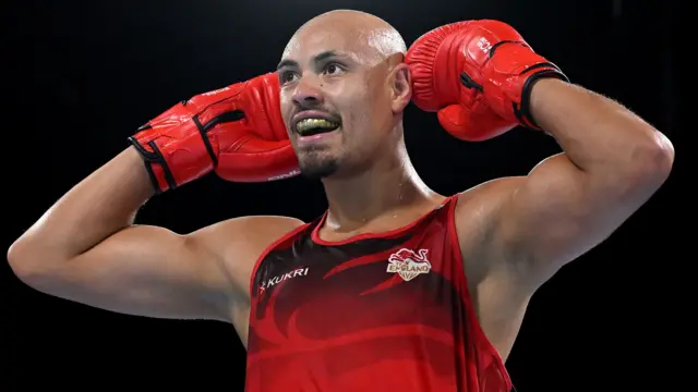 Boxer Isaac Okoh, wearing England's red vest top, holds his gloves up to his ears and smiles to celebrate his win in the 90kg semi-final