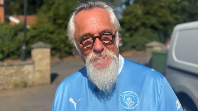 Jon Evans in Westleton. He has white hair and a long white beard. He is wearing round dark-framed glasses and a blue top.