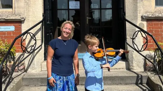 Annabel Thwaite with her son outside Framlingham College. She has blonde hair and is wearing a blue T-shirt and skirt and is smiling. Her son is playing the violin