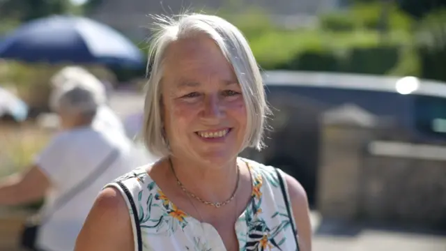 A head and shoulders shot of Gill Wood, outside Westleton Village Hall. She has bobbed white hair and is wearing a floral sleeveless top. She is smiling broadly.