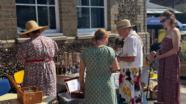 A group of four people looking at a stall outside Westleton Village Hall. They are wearing summer clothes.