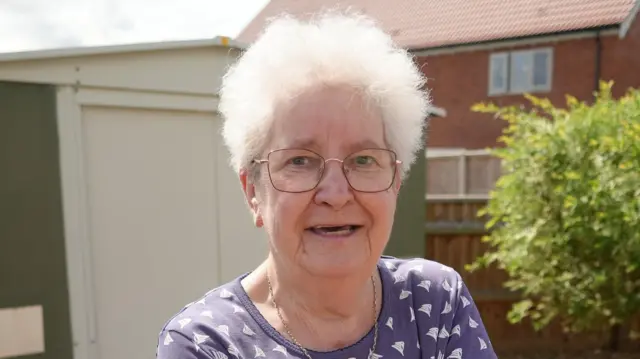 A head and shoulders image of a woman with short white hair, glasses and a blue top. She is standing in a back garden, with a fence, tree and house behind her.