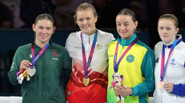 Sacha Hickey, wearing England's white and red jacket. stands on the podium with her gold medal around her neck next to Northern Ireland's Kaci Rock, wearing a green jacket and holding her silver medal