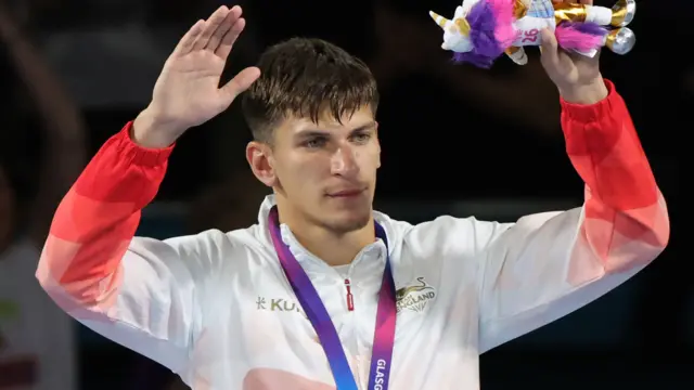 Patris Mughalzai, wearing England's white and red tracksuit jacket, raises his hands in the air with his gold medal around his neck