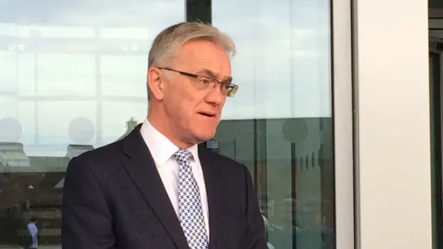 Mark Pendlington, who is standing in front of a large glass window. He has short grey hair, is wearing glasses, a dark suit, while shirt and tie.