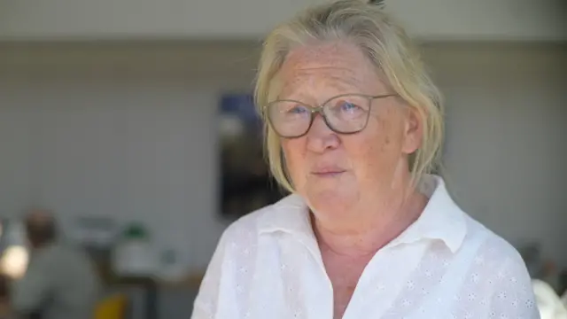 A head and shoulders shot of Barbara Buckley, inside Westleton Village Hall. She has a blonde bob and is wearing glasses and a white shirt.