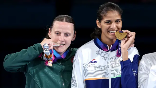 Michaela Walsh, wearing Northern Ireland's green jacket, puts her silver medal in her mouth while stading on the podium with gold medallist Jaismine Lamboria