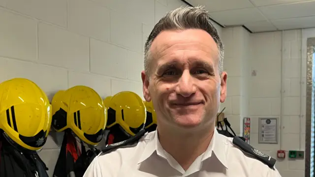 Wayne Swales has short grey hair. He is wearing a white shirt and is standing in front of a wall which has helmets and Hi-Vis jackets hung up on pegs.