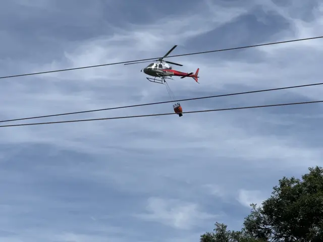 A red helicopter is in the air and flying over trees and telephone wires. It has an orange bucket attached to it at the bottom which is used to get water.