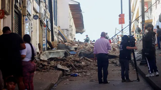 People stand around piles of rubble that have collapsed into the middle of a suburban street.