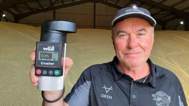Richard Beachell holds up an electronic tool used to measure moisture - its display reads 13.8%. Beachell wears a black and grey polo top with a black and grey cap.