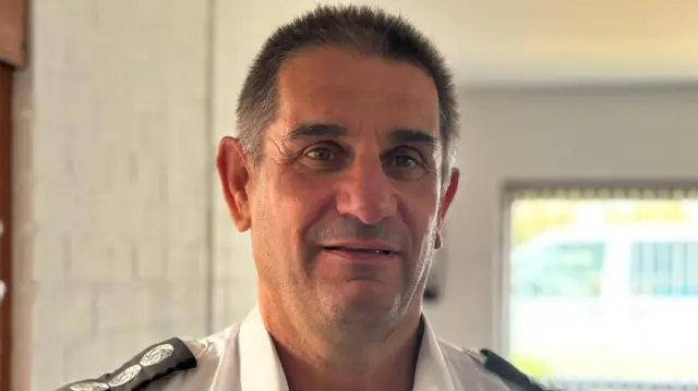 Anthony Gould is standing in a white room. There is a window behind him. Gould has short brown hair and is wearing a white shirt.