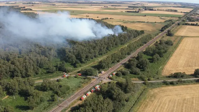 Smoke can be seen in the sky, with trees burning and red fire engines parked on the side road. The image is a drone image.