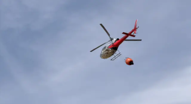 A red helicopter is in the air and has an orange bucket attached to it at the bottom which is used to get water.