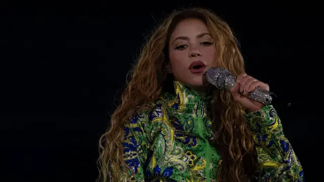 The pop singer Shakira is pictured with a microphone, wearing a colourful green, blue and yellow jacket.