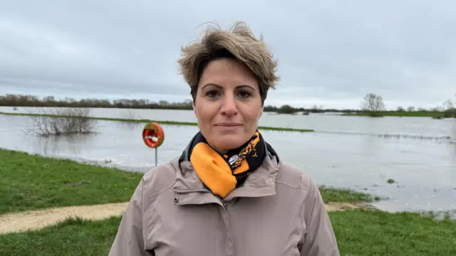 Emma Hardy poses for the camera with a body of water behind her. She wears a grey coat and an orange and black scarf.