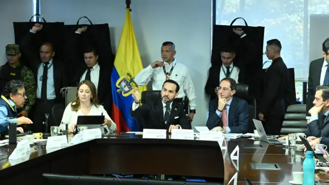 Abelardo de la Espriella gestures with his right hand as he sits at the head of a large circle of desks and people in an operations room.