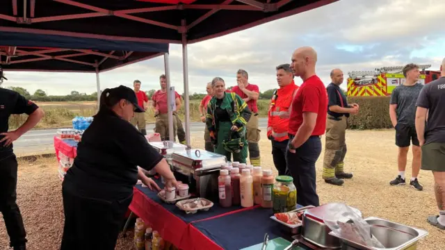 Firefighters and other emergency service personnel queue up at a covered stall serving burgers and bacon rolls. The servers are dressed in black trousers and T-shirt. A fire engine can be seen behind a hedge in the background.