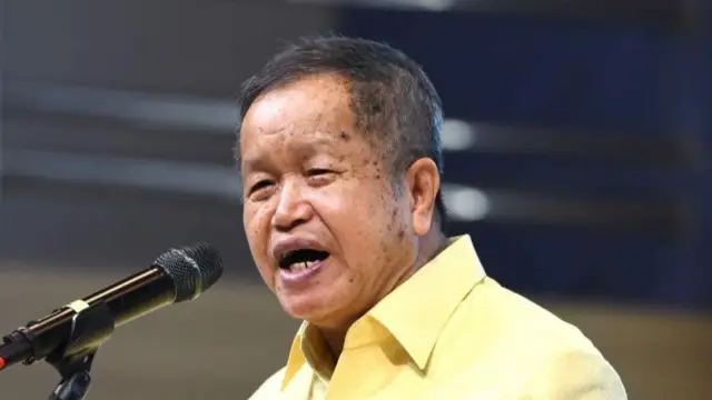 Thongchai speaking into a microphone, wearing a yellow shirt.