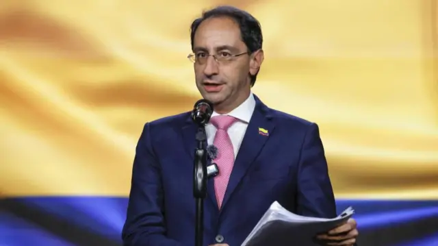 José Manuel Restrepo Abondano is pictured speaking behind a microphone holding a sheaf of papers and wearing a suit and tie. There is a large Colombian flag behind him.