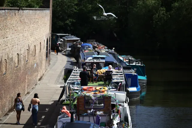 There is a canal with numerous boats moored along the edge, some people can be seen onboard. To the left is a path and high brick wall. Two women in jeans can be seen walking along the path. The sky is not visible but it is clearly sunny
