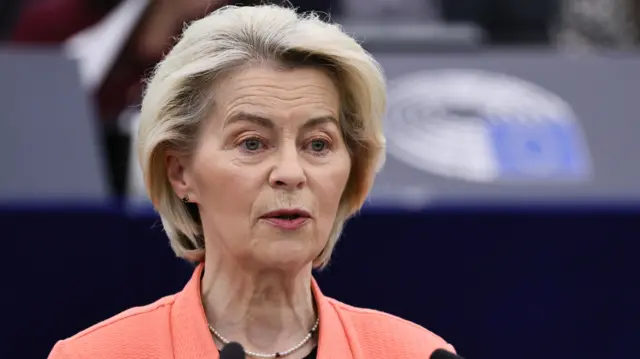 A head shot of a woman with short hair and wearing an orange jacket, who is speaking