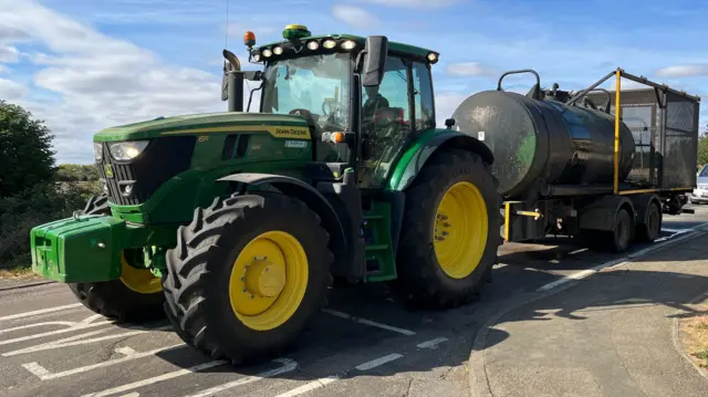 A green tractor is on a road near a junction. It is pulling a trailer which has a green water carrier. A silver car is behind the tractor.