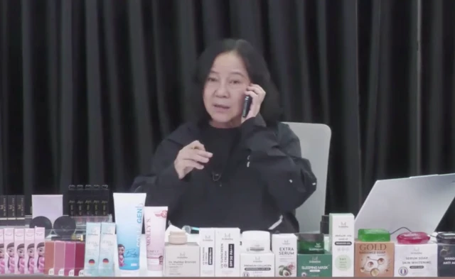 A woman wearing a black jumper sits at a table lines with packaged beauty products as she holds a phone to her ear, pulling a surprised facial expression