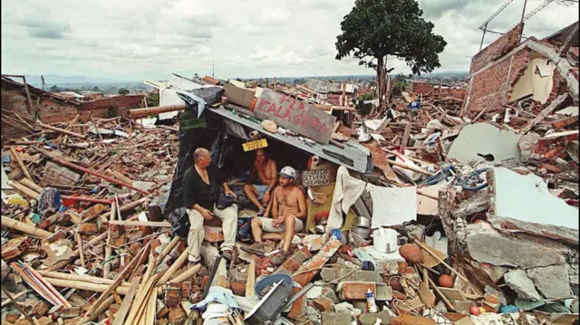 Three men sit in a makeshift shelter amid a wide slew of rubble and debris.
