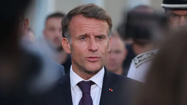 Emmanuel Macron is showing wearing a suit and tie with a serious expression on his face.