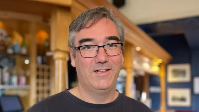 Mark Whittington has short grey hair. He is wearing black framed glasses and a black T-shirt. He is standing in front of a bar which is made out of wood.