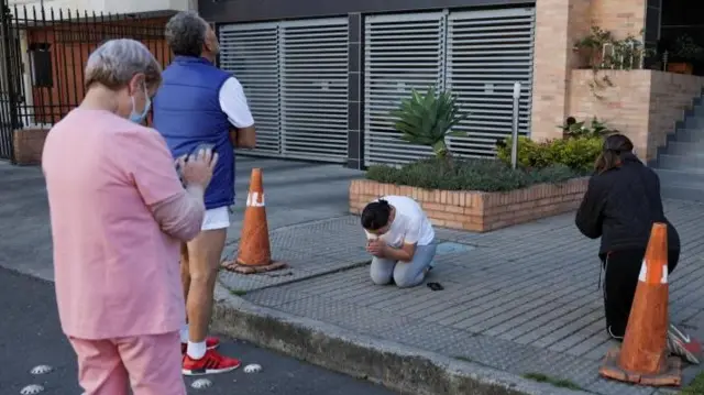 A woman is shown kneeling in prayer on the pavement outside an apartment building. Other people are standing nearby looking up at the building, which is out of view.