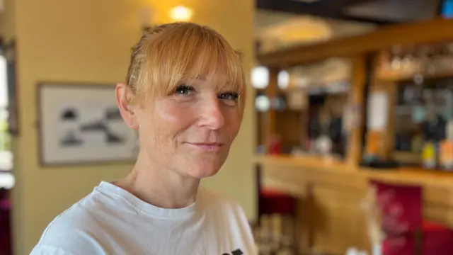 Dawn England has blonde hair in a ponytail and a long fringe. She is wearing a white T-shirt and is standing inside a pub. There is a wooden bar on the right.