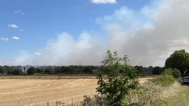 A large grey plume of smoke over the dry fields, under a blue sky.