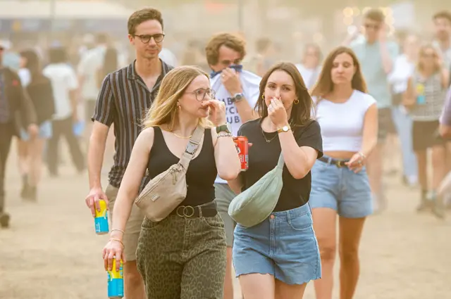 Festival goers cover their faces from the dust - three girls and two boys walk on a dusty field in London.