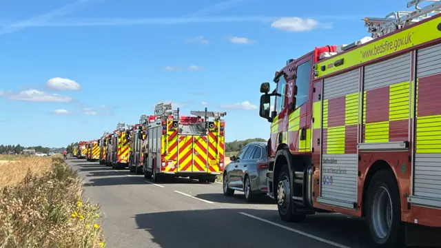 A line of fire engines on the road next to a field.