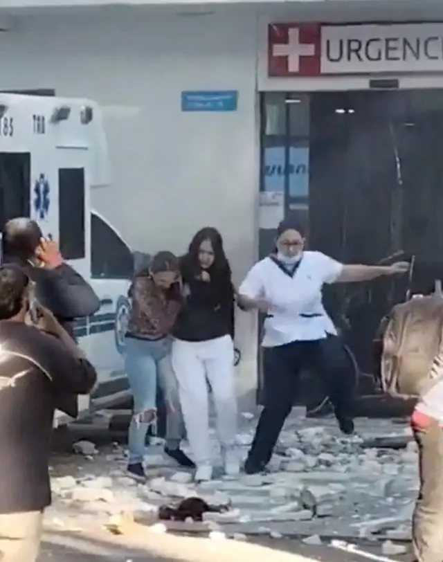 People are shown clambering through piles of rubble outside a hospital, beside a parked ambulance.