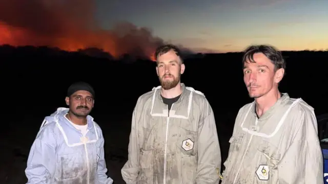 Three men stand in the dark with beekeeping suits. Flames can be seen behind them.