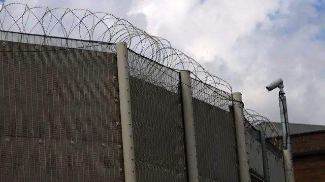An image of outside of a prison, it shows its walls and razor wire at the top