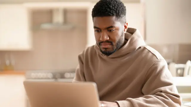 A young person in a beige hoodie working on a laptop at a table in a softly lit home kitchen. Cabinets and appliances are blurred in the background, with the focus on the computer screen and workspace.