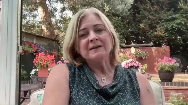 Debbie Adlam wears a green top and has blonde hair, she sits in a garden