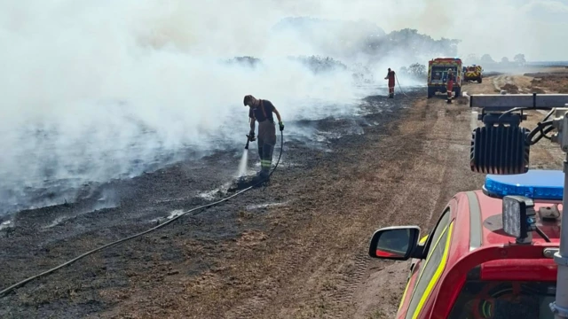 A firefighter tackling a hotspot on a heathland with a hose. There is another firefighter in the distance behind him with several vehicles, and a lot of smoke.